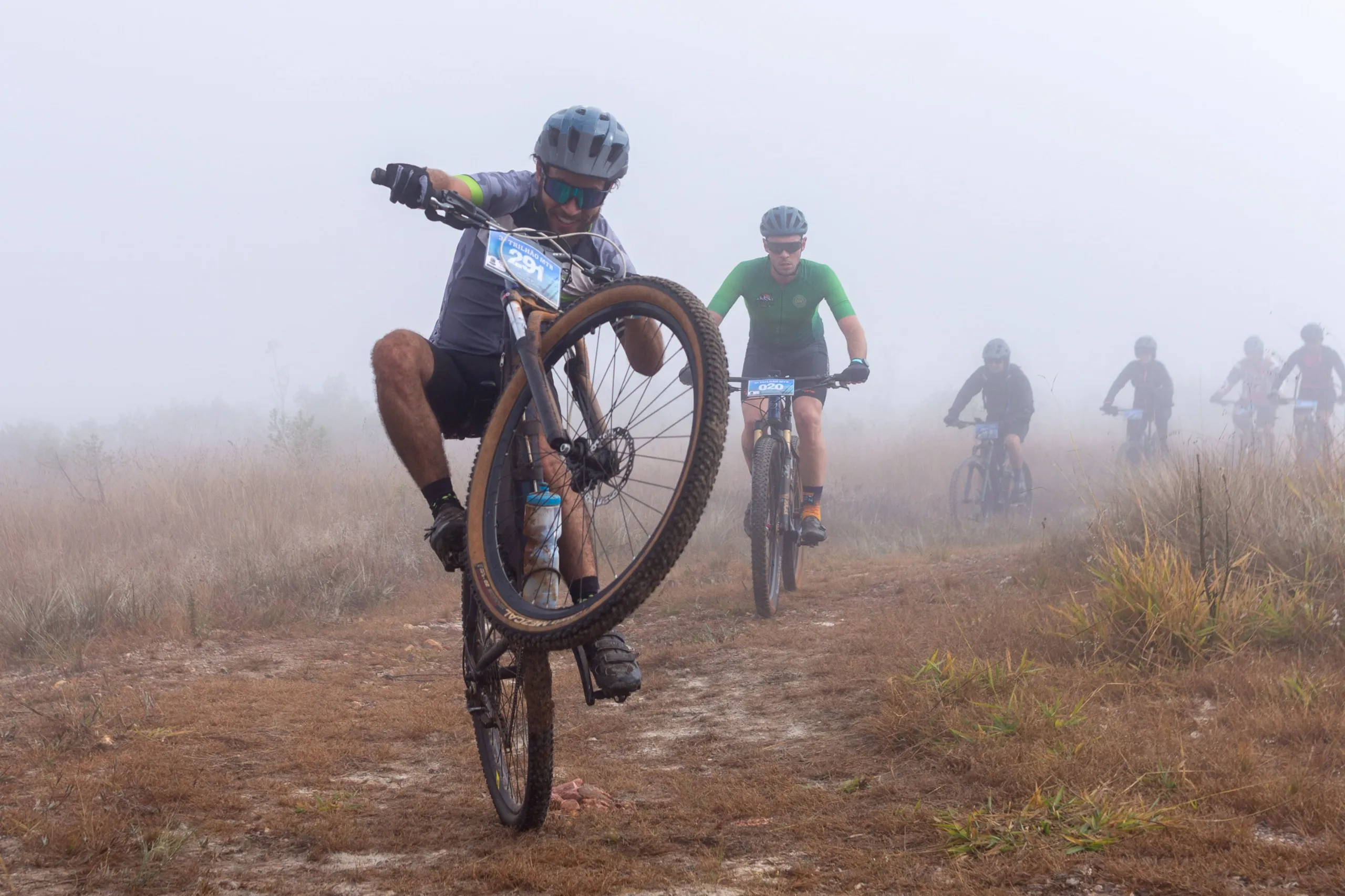 Atleta fazendo wheelie na frente do grupo em estrada de terra coberta de névoa
